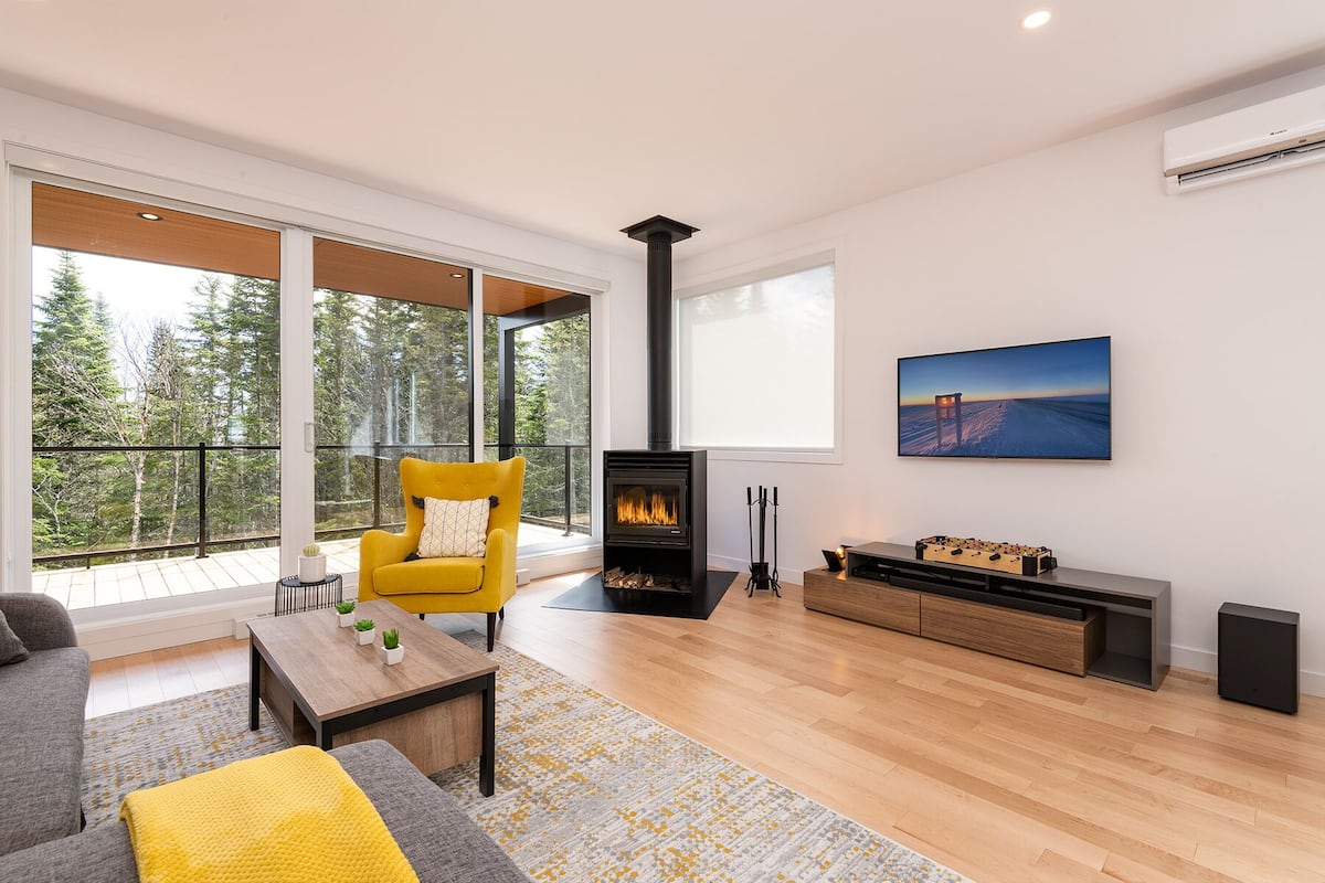 A modern living room with a fireplace, a sofa, a yellow armchair, and a large window overlooking a forest.