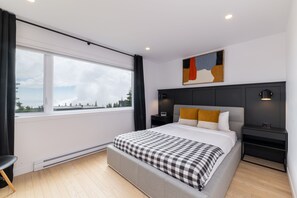 A modern bedroom with a large window, a bed with a checkered blanket, a nightstand, and a wall-mounted lamp. - Les Chalets Forêt (Petite-Riviere-Saint-Francois)