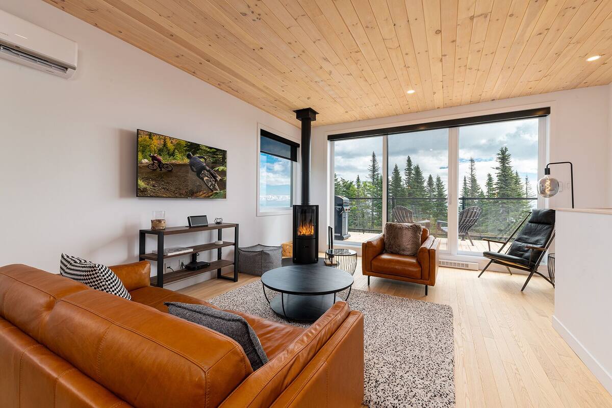 A modern living room with a leather sofa, a black fireplace, a dining table, and a view of a balcony with a grill.