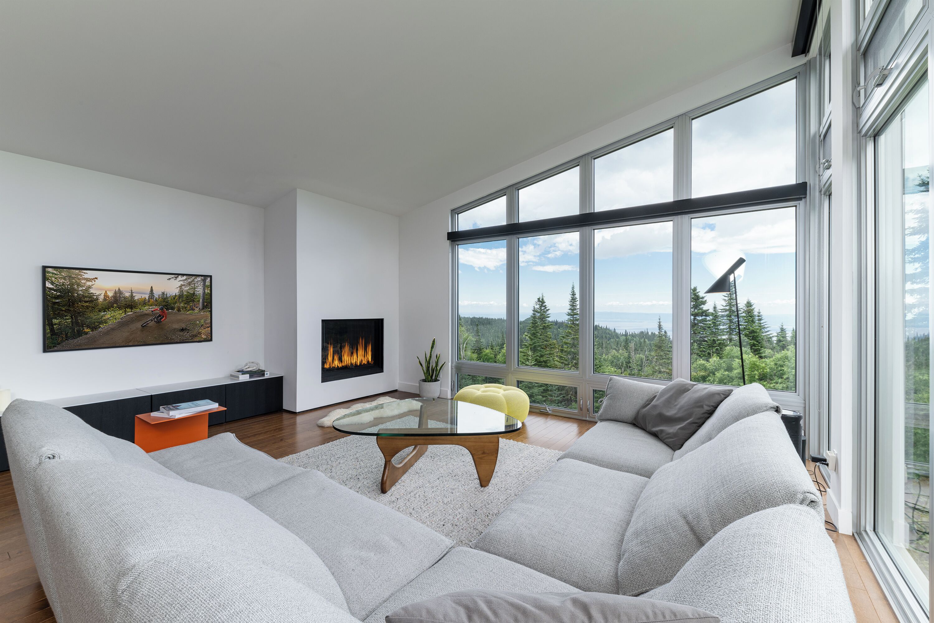 A modern living room with a large sectional sofa, a glass coffee table, a fireplace, and a large window offering a view of trees and mountains.