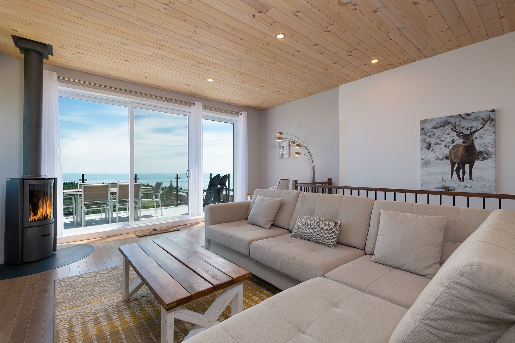A modern living room with a large sofa, a wooden coffee table, a fireplace, and a large window offering a view of the outdoors.