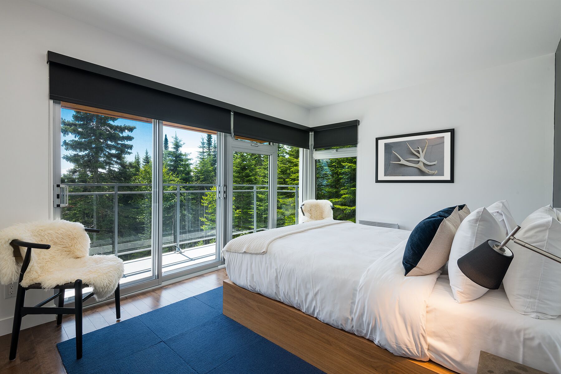 A modern bedroom with a large bed, a white chair, and a view of trees through floor-to-ceiling windows.