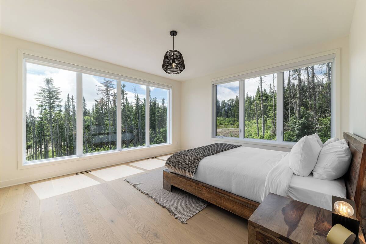 A bedroom with a large window, a wooden bed, a bedside table, and a hanging light fixture.