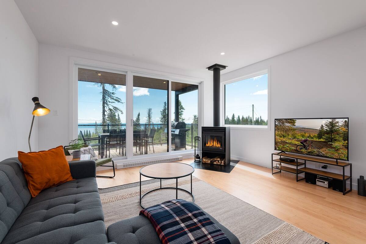 A modern living room with a sofa, a fireplace, a TV, and a balcony with a view of the forest and a body of water.
