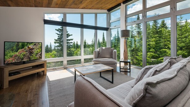 A modern living room with a large TV, comfortable seating, and expansive windows offering a view of a forested area. - Les Chalets Forêt (Petite-Riviere-Saint-Francois)