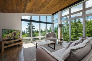 Una sala de estar moderna con un televisor grande, asientos cómodos y amplias ventanas que dan a una zona boscosa.
