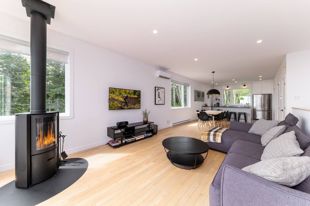 A modern living room with a large black wood-burning stove, a grey sectional sofa, and a dining area with a kitchen in the background.