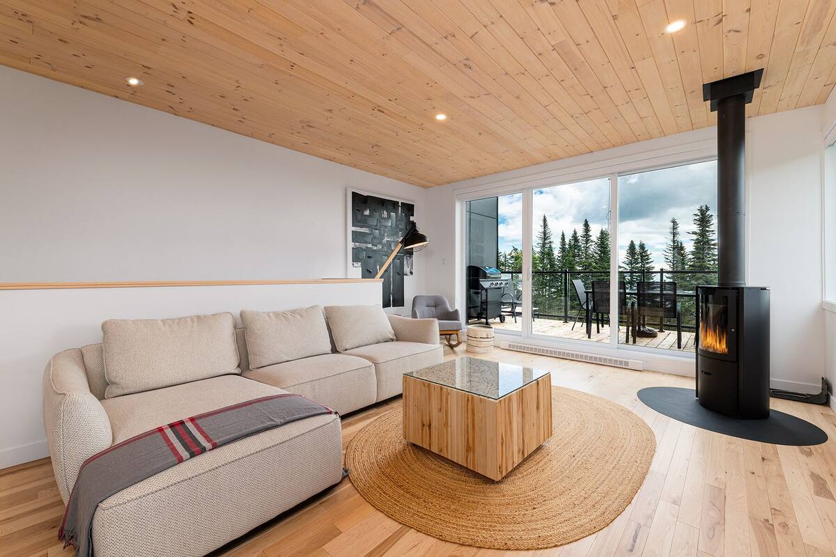 A modern living room with a wooden ceiling, a glass sliding door leading to a balcony with a view of trees, a black fireplace, and a beige sofa.