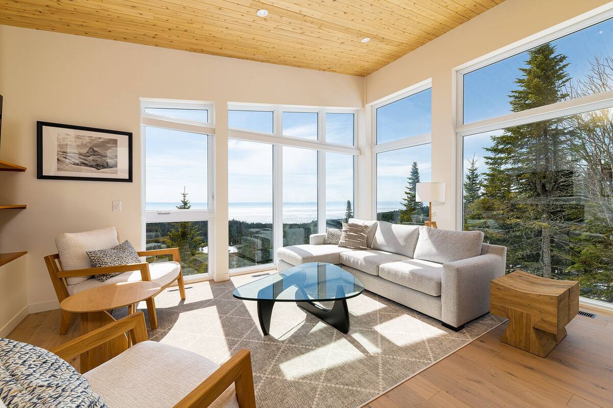 A spacious living room with a large window offering a view of a forest and mountains. There is a sofa, a coffee table, and a chair with a rug.