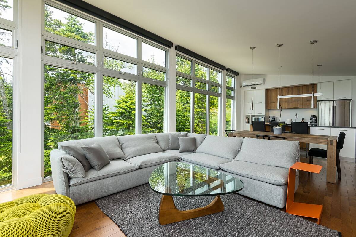A modern living room with a sectional sofa, a glass coffee table, and large windows offering a view of greenery.