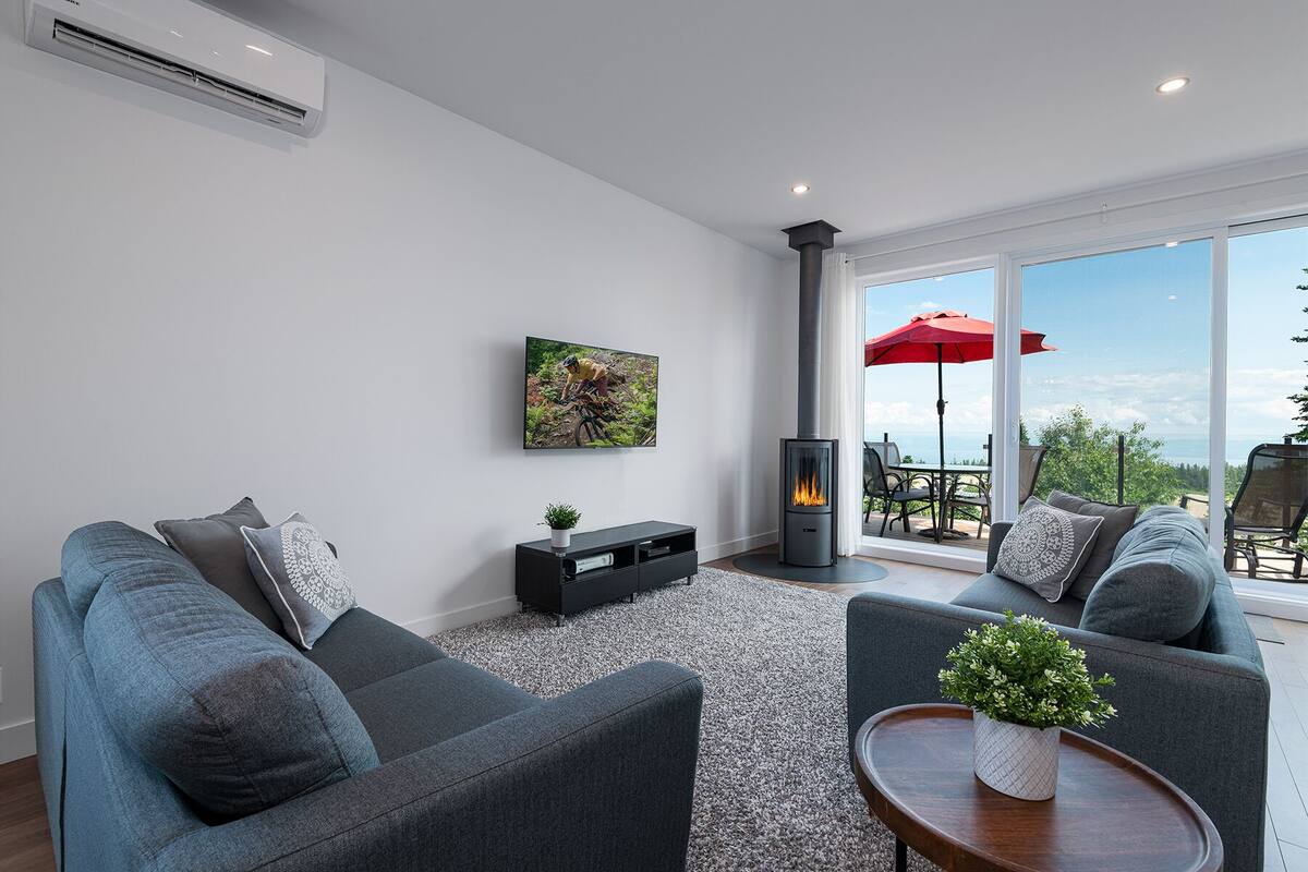 A modern living room with grey sofas, a flat-screen TV, a fireplace, and a large window overlooking a scenic view.