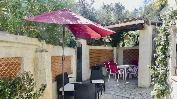 Een terras buiten met stenen vloer, rieten meubels en een rode parasol.