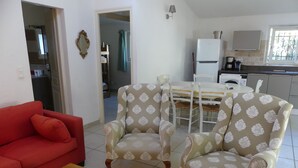 Een woonkamer met een rode bank, twee gestoffeerde fauteuils, een houten salontafel, een eettafel met stoelen, een keukenhoek met een koelkast, en een raam met uitzicht.