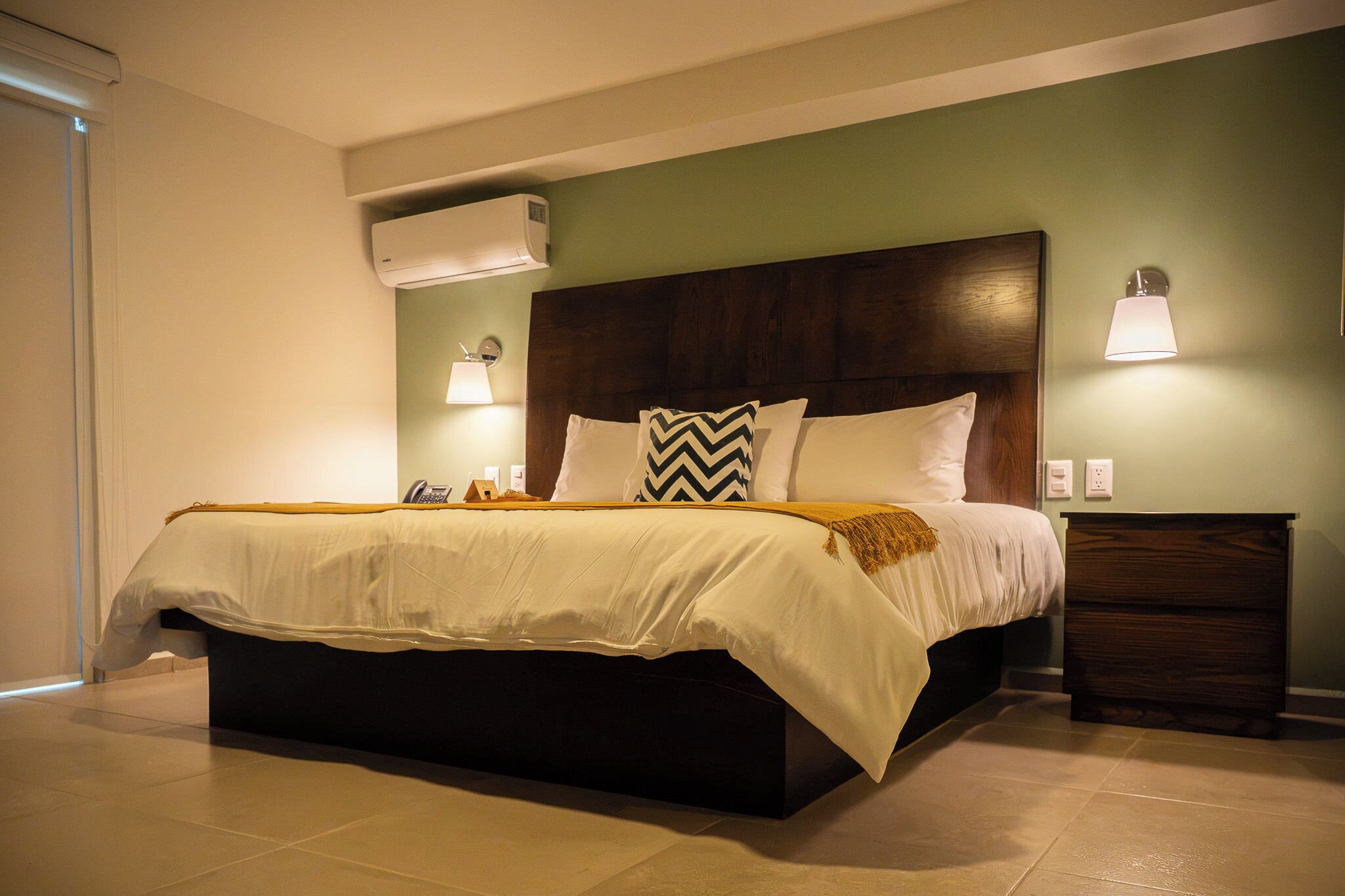 A bedroom with a bed, bedside lamps, a nightstand, and an air conditioning unit.