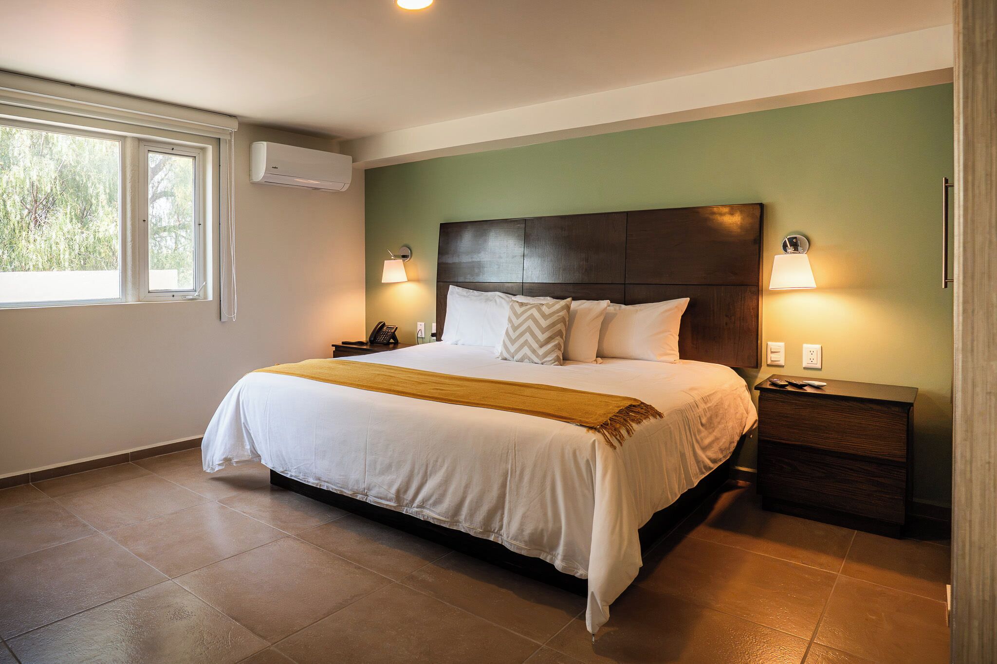 Cama bem arrumada com cabeceira, abajures, mesa de cabeceira e janela com persianas.