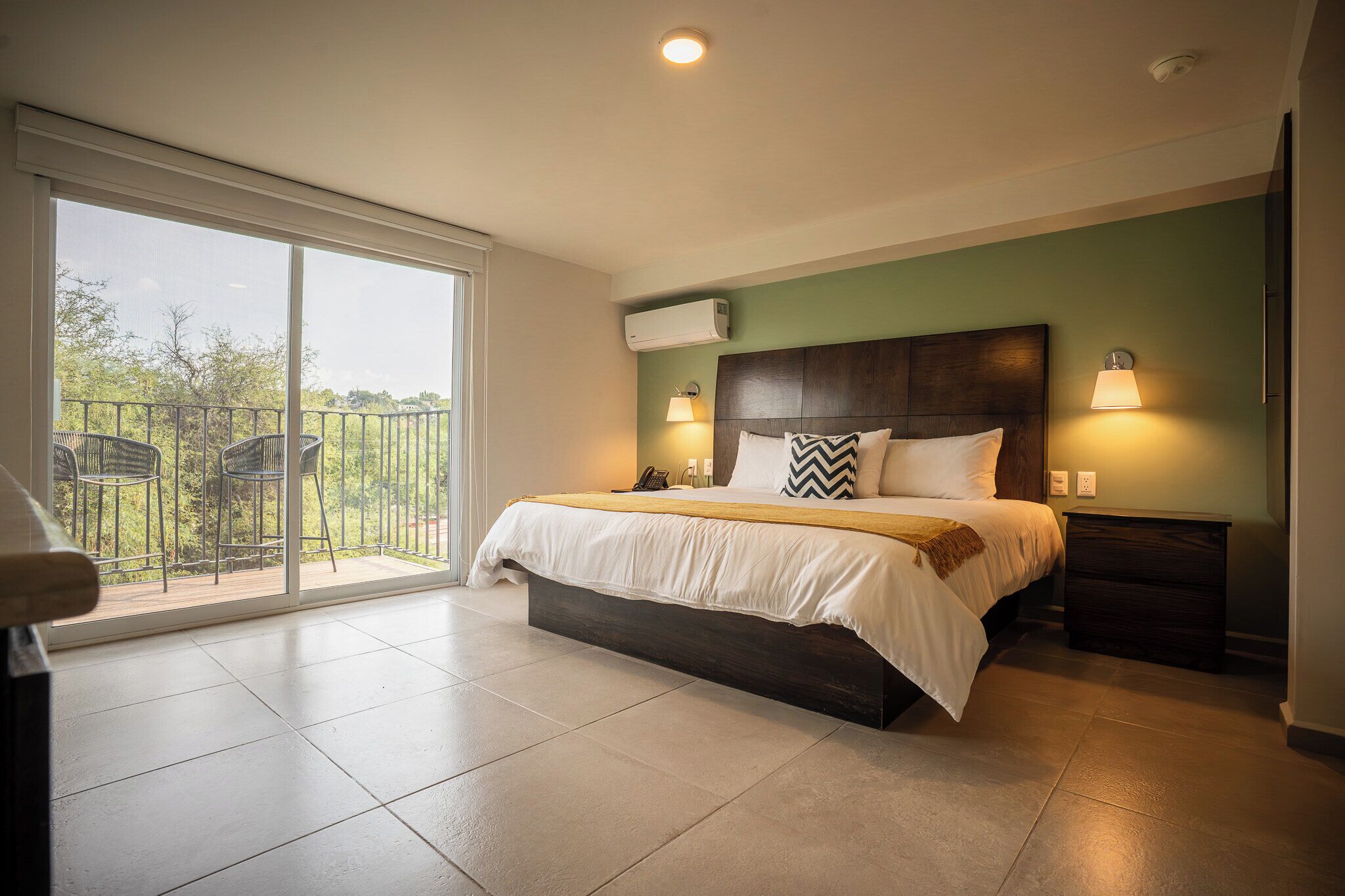 A bedroom with a large bed, two bedside lamps, a nightstand, a sliding glass door to a balcony, and an air conditioning unit.