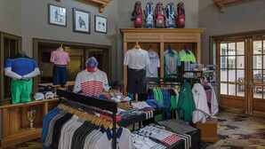 Ein Bekleidungsgeschäft mit Kleiderstangen voller Hemden und Hosen, einer Schaufensterpuppe und ausgestellten Golftaschen.