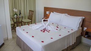 A neatly made bed with white linens and a heart-shaped towel arrangement, a small table with a bottle and flowers, and a chair in a hotel room. - Pousada Laranja Durigon  (Natal)