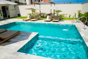 A swimming pool with lounge chairs and umbrellas, surrounded by a tiled patio and greenery.
