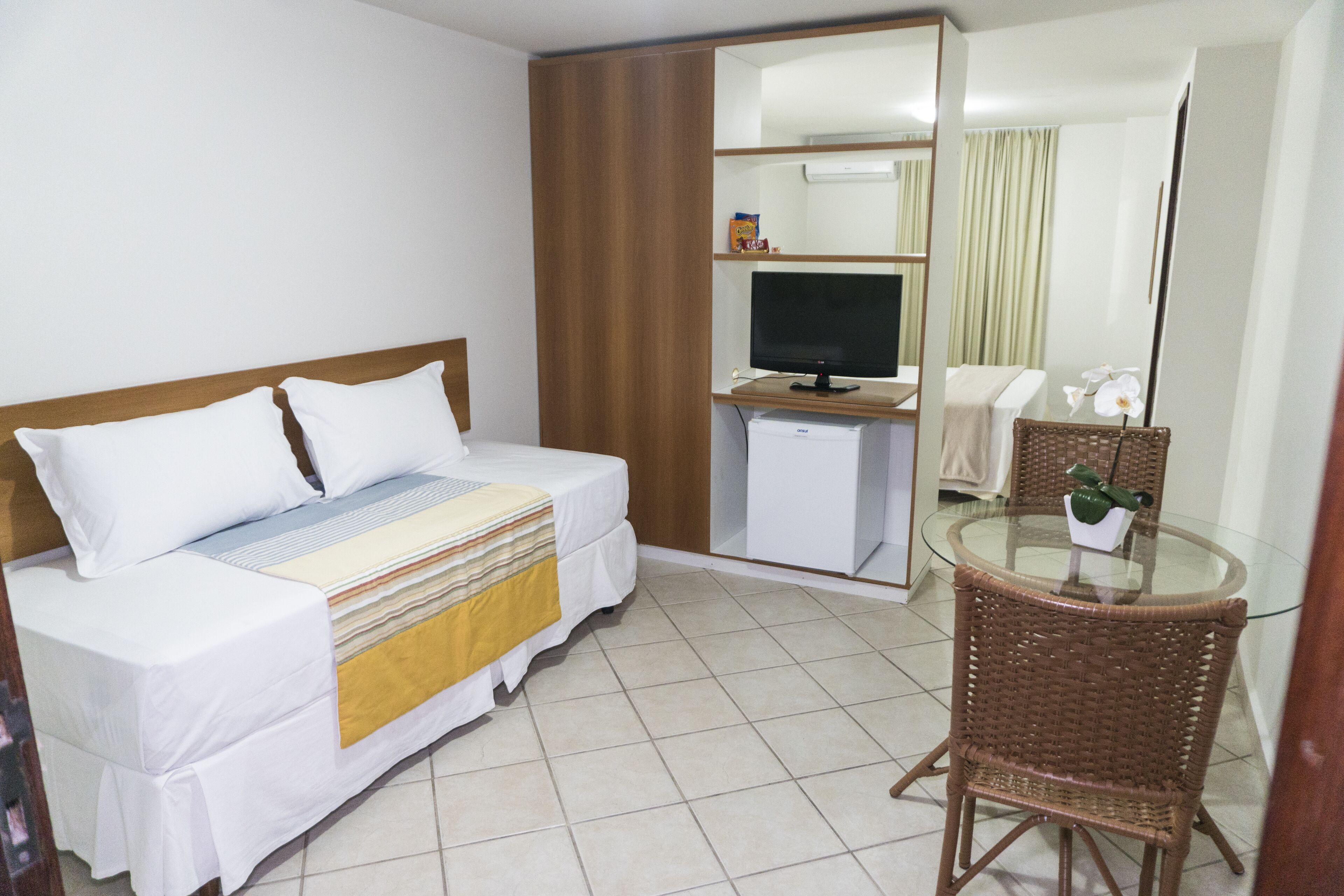 A hotel room with a bed, a desk with a computer, a chair, and a small table with a flower vase.