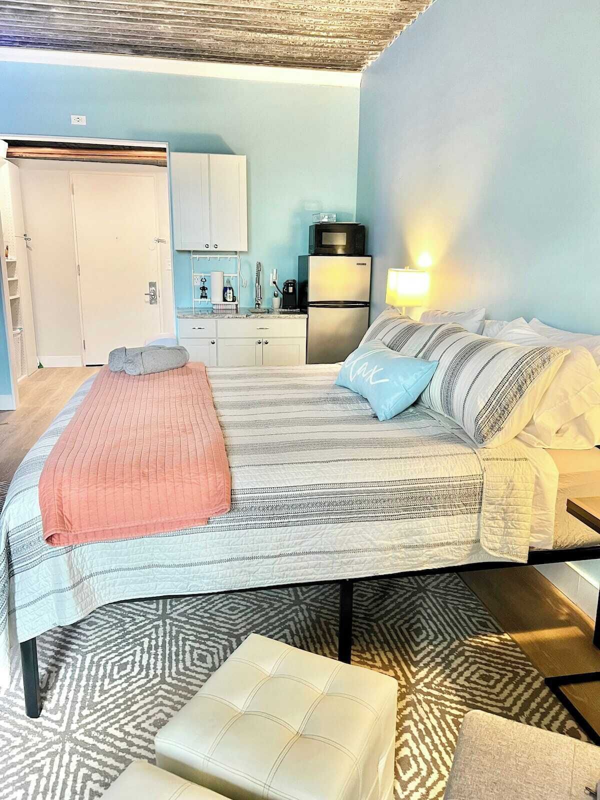 Een slaapkamer met een bed, nachtkastje, lamp, kast en een kitchenette.