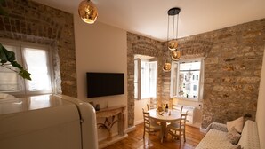 A cozy living room with a stone wall, wooden floor, a sofa, a dining table with chairs, and hanging pendant lights.