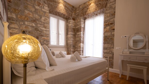 A bedroom with a stone wall, a bed with white linens, a bedside table, a lamp, and a dresser with a mirror.