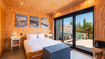 Une chambre à coucher en bois, dotée d’un grand lit, de deux tables de chevet, d’un banc et offrant une vue sur une terrasse aménagée avec du mobilier d’extérieur.