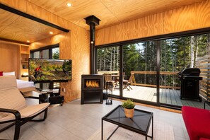 A modern living room with a large TV, a fireplace, and a view of a forested area. - Les Refuges (Petite-Riviere-Saint-Francois)