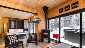 A cozy room with a bed, a chair, a table, a fireplace, and large windows with a snowy view. - Les Refuges (Petite-Riviere-Saint-Francois)