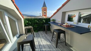 Un balcón con suelo de madera, dos taburetes, una mesa pequeña con una tabla de quesos y copas de vino, y vistas a una torre de iglesia.