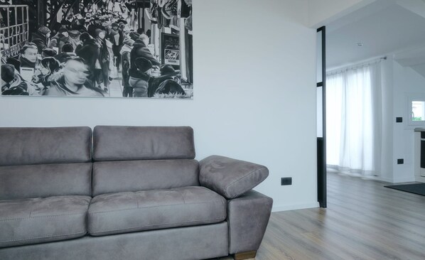 A modern living room with a grey sofa, a large black-and-white photograph on the wall, and a view of a bright hallway. - Studio D'artista (Chiavari)