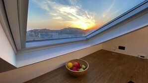 Una habitación con tragaluz que da a una puesta de sol, una mesa de madera con fruteros y una pared blanca.