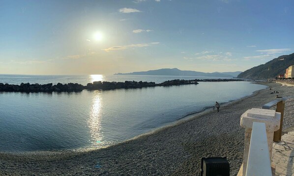 A pebbled beach with a rocky breakwater, a clear sky, and distant mountains.