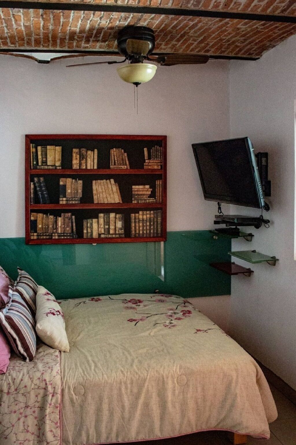 A bedroom with a green accent wall, a wooden bookshelf filled with books, a television mounted on the wall, and a bed with floral bedding.