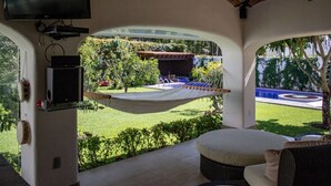 Een overdekt terras met een hangmat, een aan de muur bevestigde televisie en uitzicht op een zwembad en tuin.