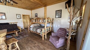 Een knus houten interieur met een houten plafond, een bank, een bed met een gebreid kleed, een paarse fauteuil en een houten boekenkast.