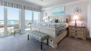 Une chambre à coucher comprenant un lit, un banc, une table de chevet et de grandes fenêtres donnant sur la plage.