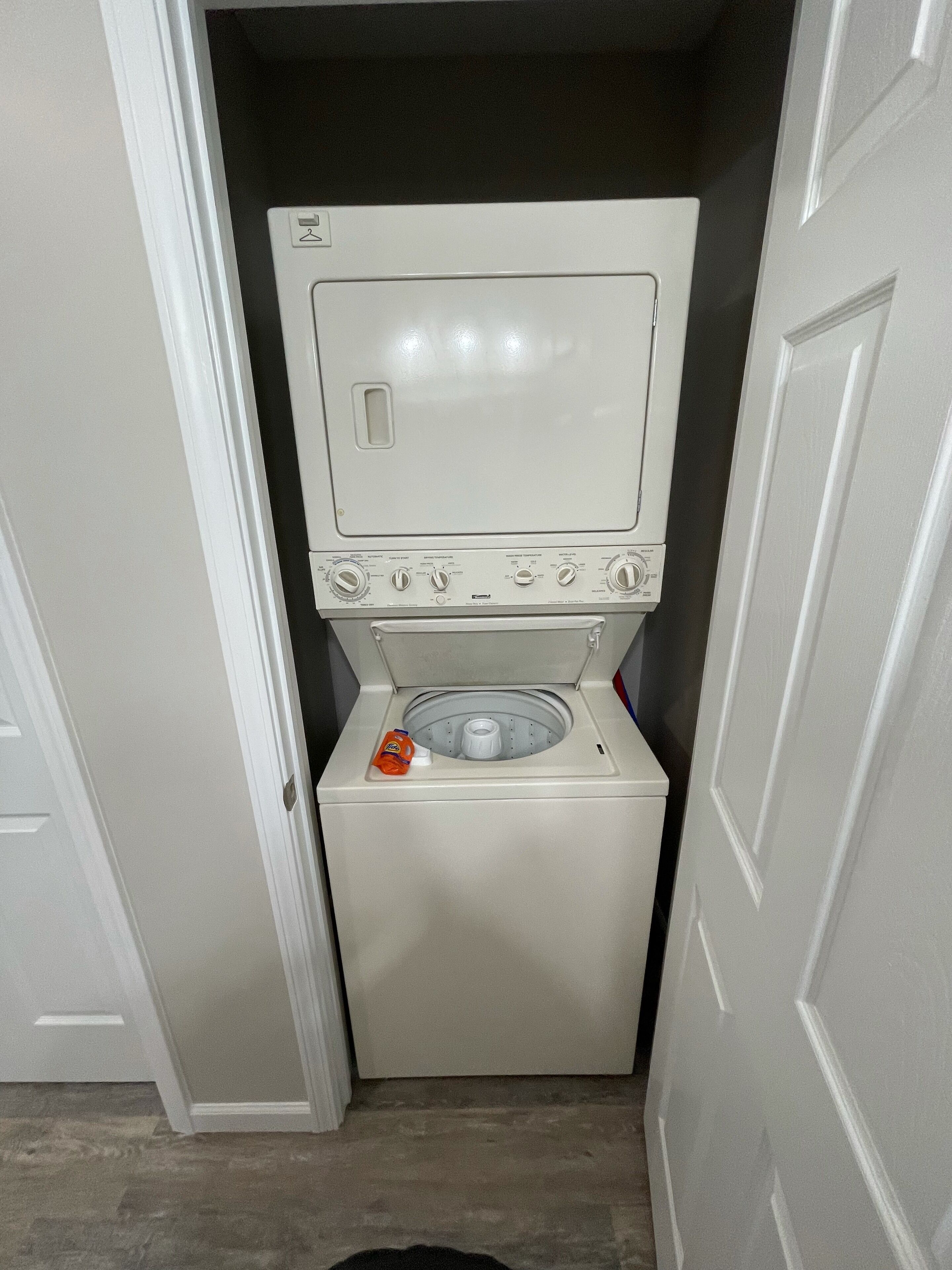 Un appareil compact, blanc, combinant lave-linge et sèche-linge, installé dans un couloir étroit, avec une porte d’un côté et un mur de l’autre.