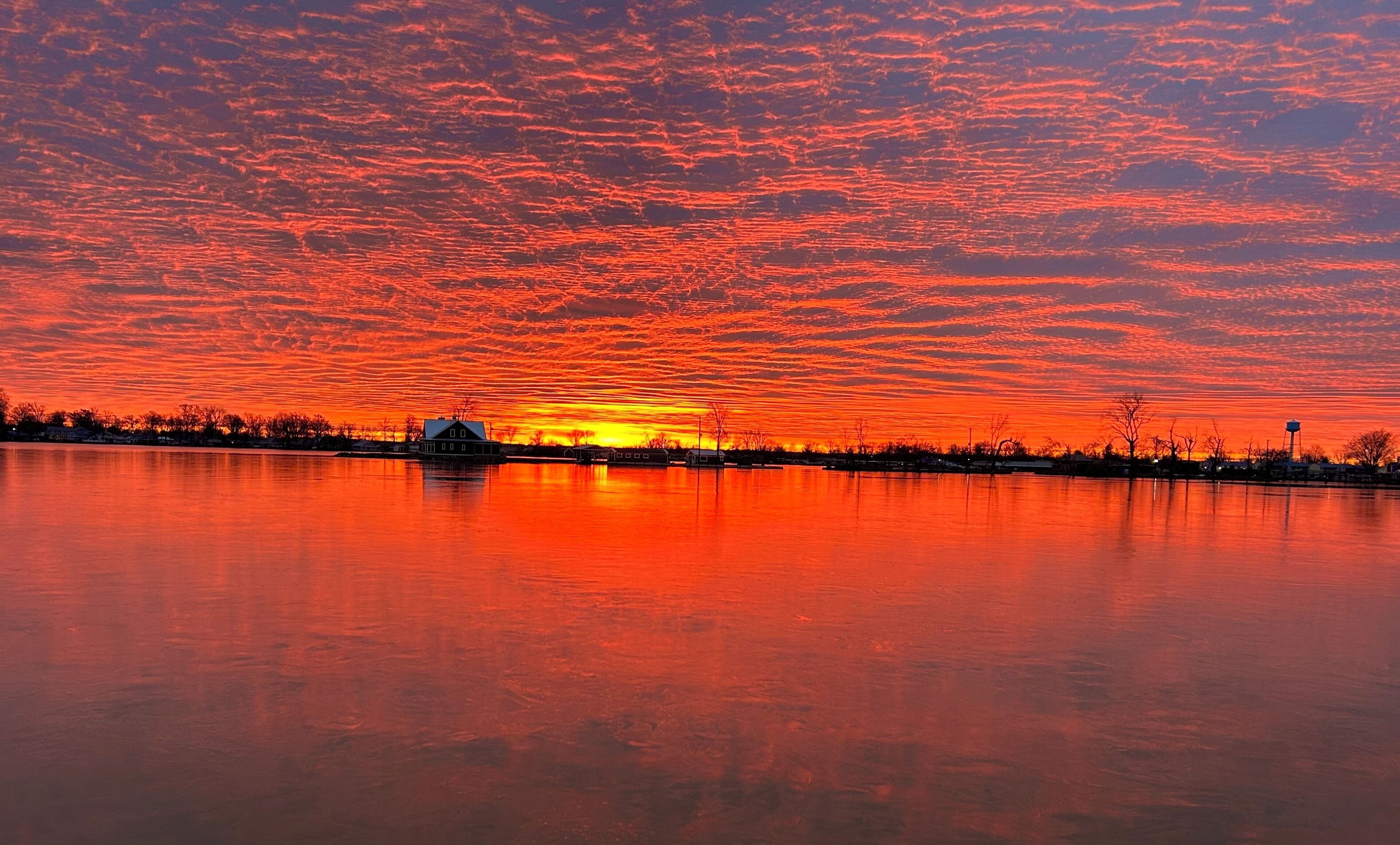 Un atardecer sobre un lago tranquilo con una casa y árboles silueteados contra el cielo.