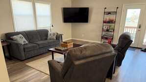 A living room with a grey sofa, a brown leather chair, a coffee table with a chess set, a TV mounted on the wall, and a sliding glass door leading outside.