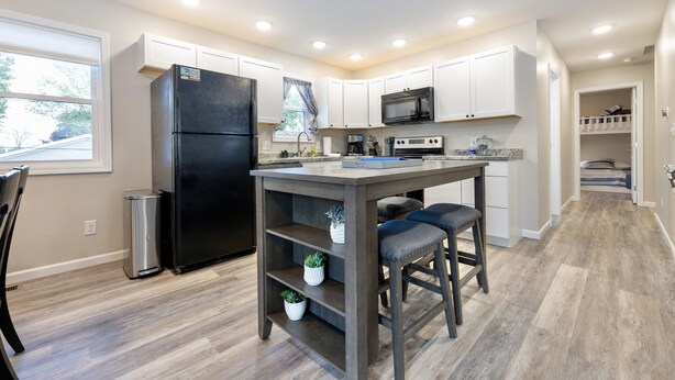 A modern kitchen with white cabinets, a black refrigerator, a microwave, and a coffee maker. There is a small island with stools and potted plants.