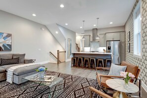 Una sala moderna con sofá, mesa de centro y alfombra estampada. Se ve una cocina con taburetes de bar y un refrigerador al fondo.