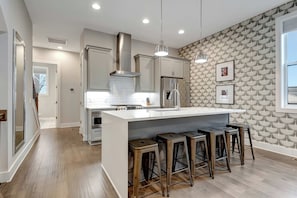 A modern kitchen with a central island, bar stools, stainless steel appliances, and a patterned wall.
