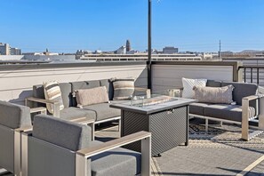 Un espacio al aire libre con zona de estar, chimenea decorativa, alfombra estampada y vista a la ciudad de fondo.