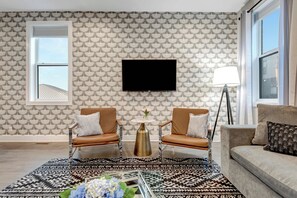 Un salón con papel tapiz estampado, un televisor montado, dos sillones, una mesa de centro y una lámpara de pie.