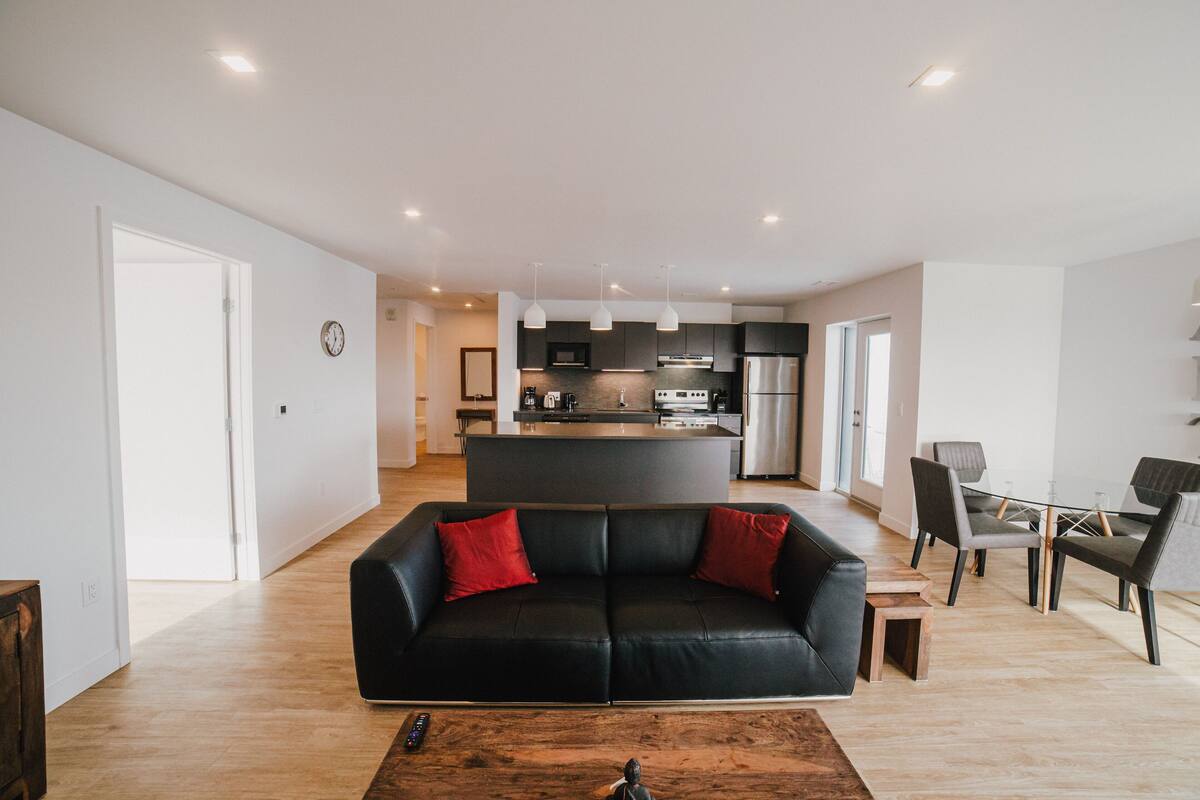 A modern living room with a black leather sofa, wooden coffee table, and a dining area with chairs and a glass table.
