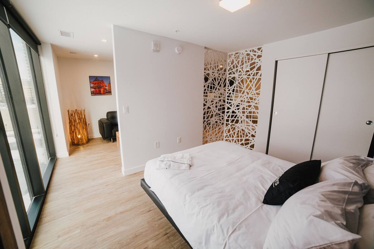 A modern hotel room with a large bed, a decorative wall panel, and a view of the city through large windows.