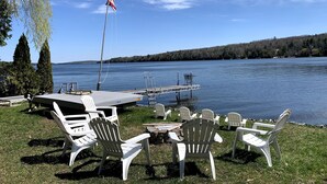 A lakeside deck with white chairs and a table, overlooking a calm lake with a dock and an American flag. - on gorgeous Great Pond (lake), great for families!  (Belgrade)