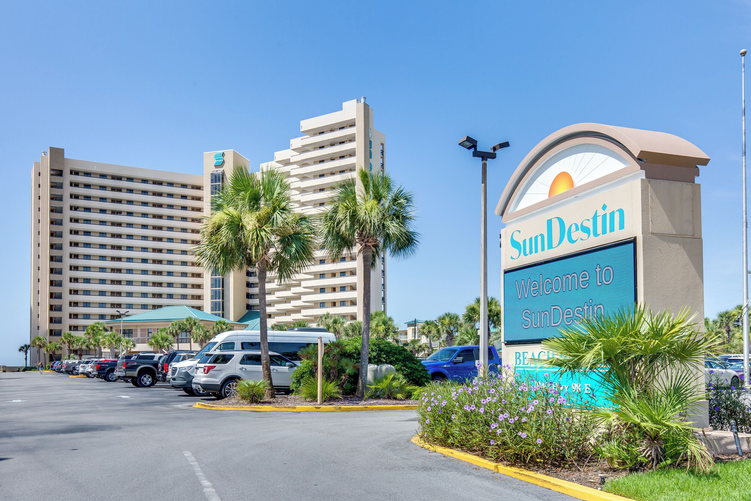 A hotel entrance with a large sign, parking lot, and a tall building in the background.